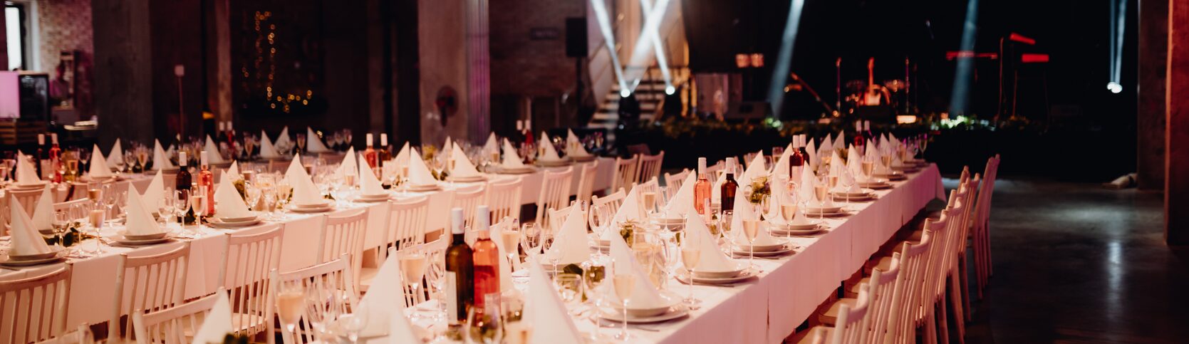 Event na kľúč - elegantne prestreté svadobné stoly s bielymi obrusmi, servírovaným vínom a pohármi pripravené na svadobnú hostinu s pódiom v pozadí.