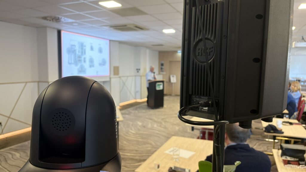 Technické zabezpečenie konferencie – kamera, reproduktor, projekčné plátno a rečník na pódiu